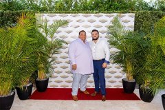 Gus and Joander smile together at camera, while standing on red carpet in front of decorative logo banner